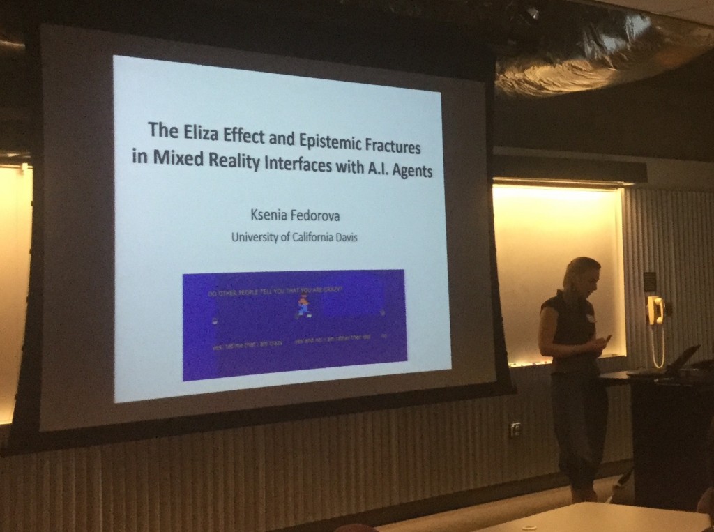 A blonde woman wearing black stands to the right of a slide that reads "The Eliza Effect and Epistemic Fractures in Mixed Reality Interfaces with A.I. Agents," Ksenia Fedorova University of California Davis