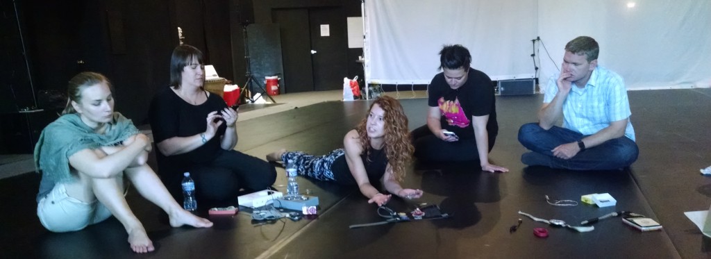 Five individuals sit on the floor with electronic components around them. The white woman in the middle is laying down and speaking to the rest.