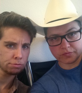 A selfie of two white masculine individuals. The person on the left has a beard, and the person on the right is wearing glasses and a cowboy hat. They are making serious travel faces.